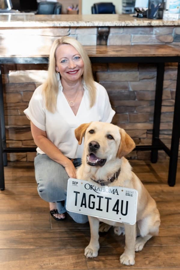 Tamara, the owner, crouching beside Archie in the office while he holds an Oklahoma plate reading TAGIT4U